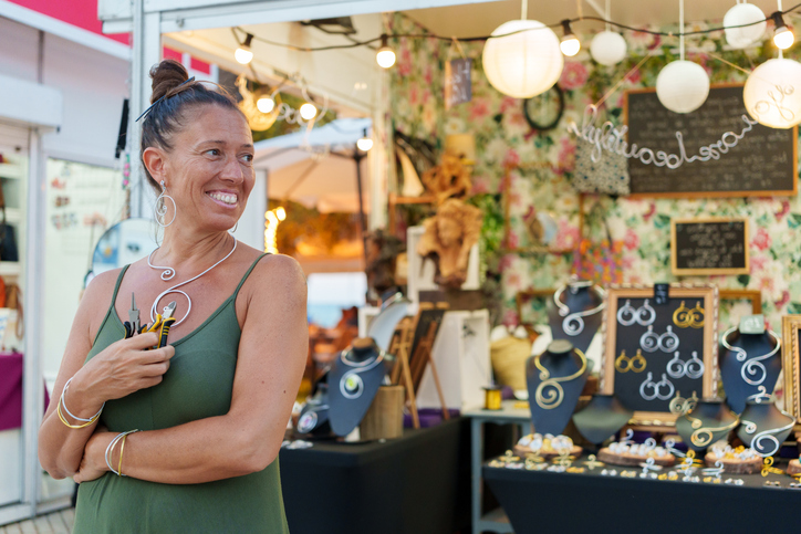 Smiling jewelry maker holding her tools, showing her handmade necklaces and earrings at her market stall