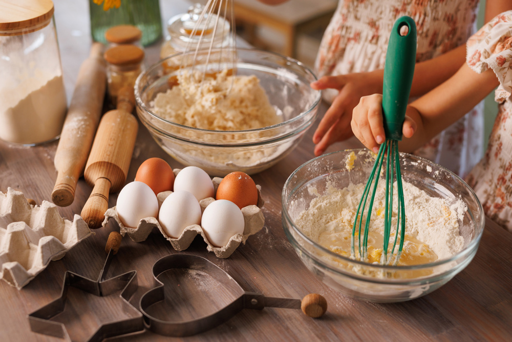 Children whisking flour and eggs in glass bowls while baking cookies together in a warm, rustic kitchen setting