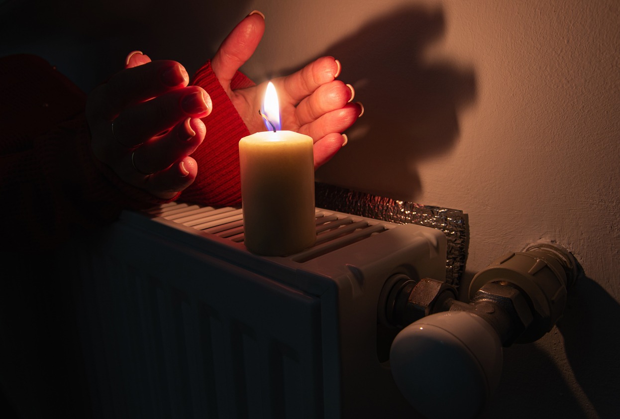 A burning candle with women's hands frozen from frost on the radiator heating in dark home. Shutdown of heating and electricity, power outage, blackout, load shedding or energy crisis, symbolic image