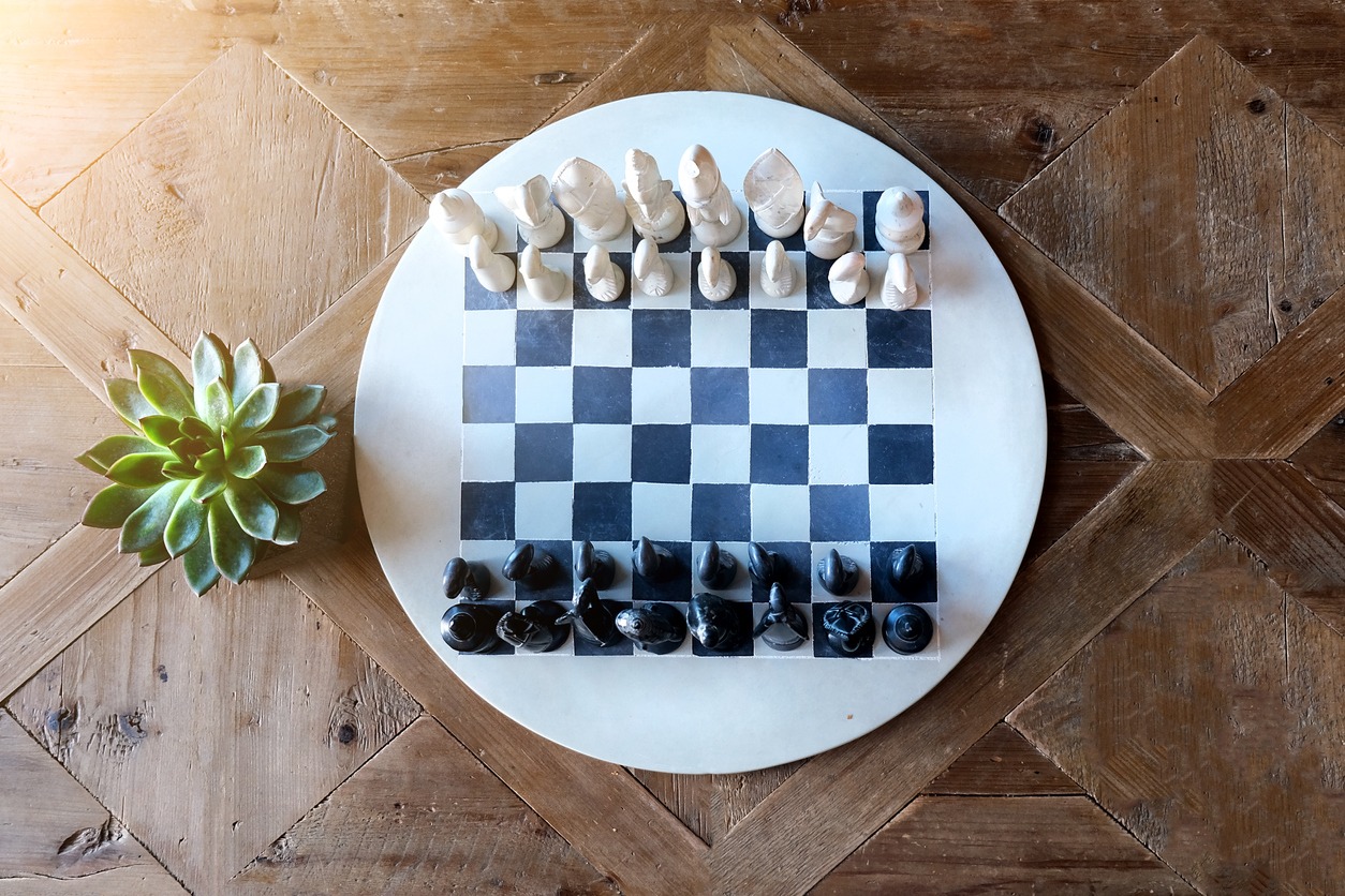 Round chessboard on a brown wooden table with green plant on a side