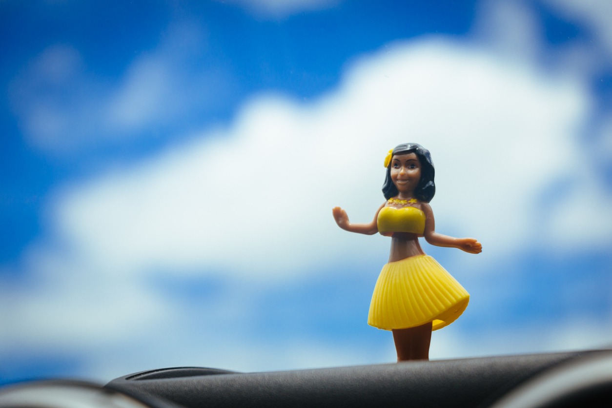 Hawaiian hula dancer doll dancing over the front panel of a car, with blue sky with some clouds on the road.
