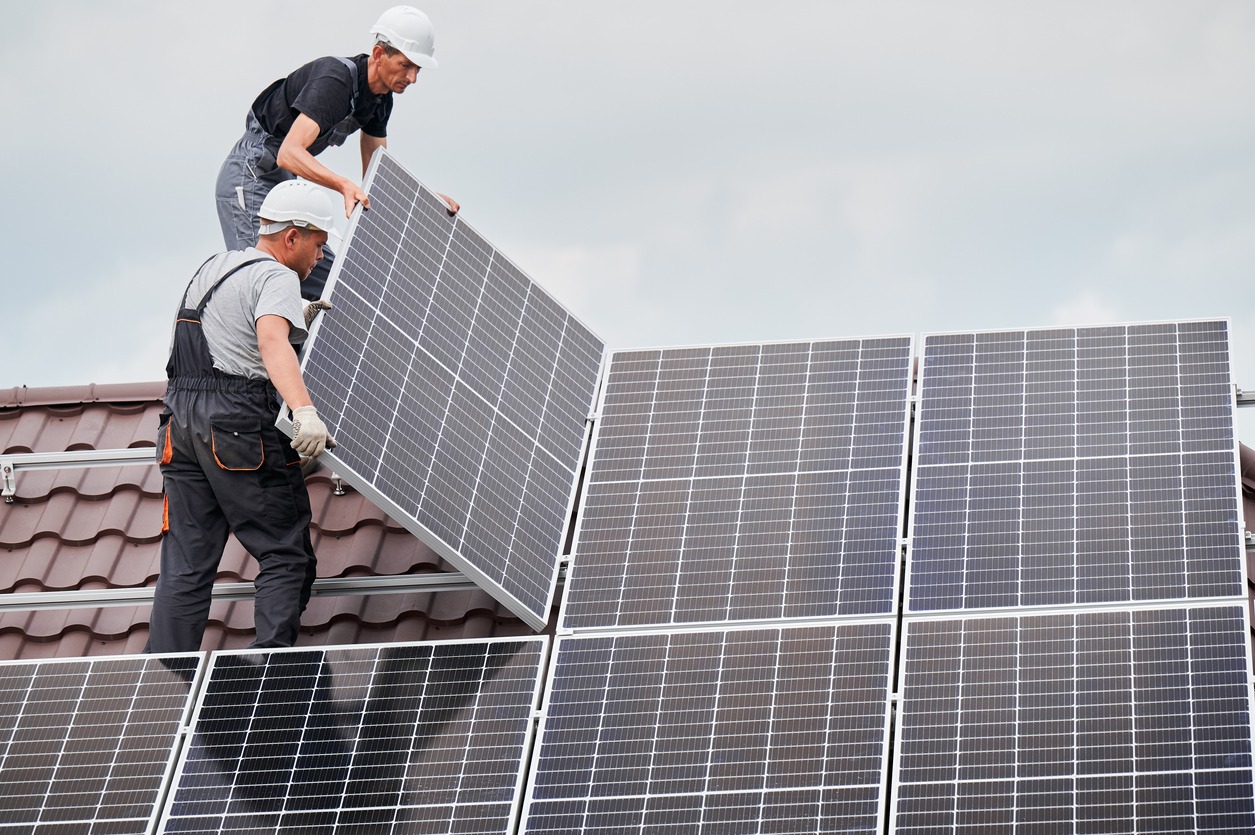 Men technicians mounting photovoltaic solar modules on the roof of the house. Builders in helmets installing solar panel systems outdoors. Concept of alternative and renewable energy