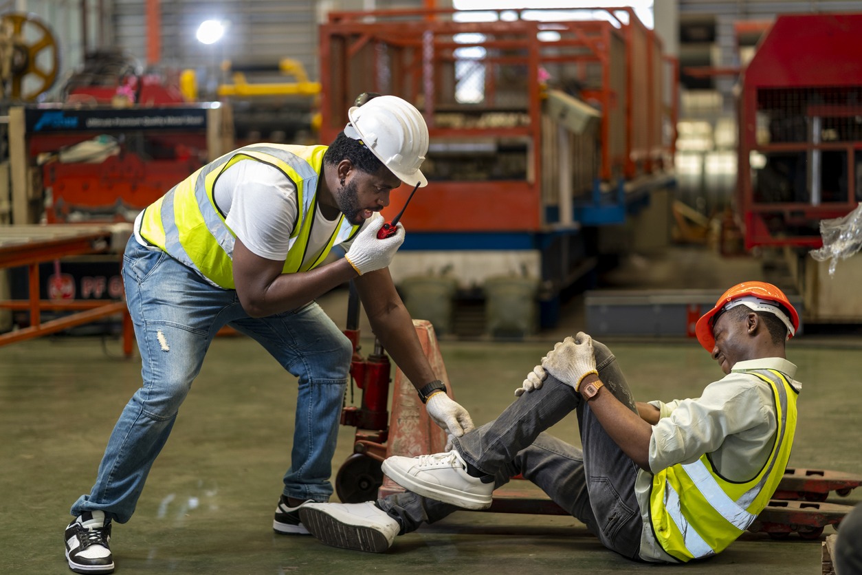 African American factory worker having accident while working in manufacturing site while his colleague is asking for first aid emergency team using walkie talkie radio for safety workplace concept