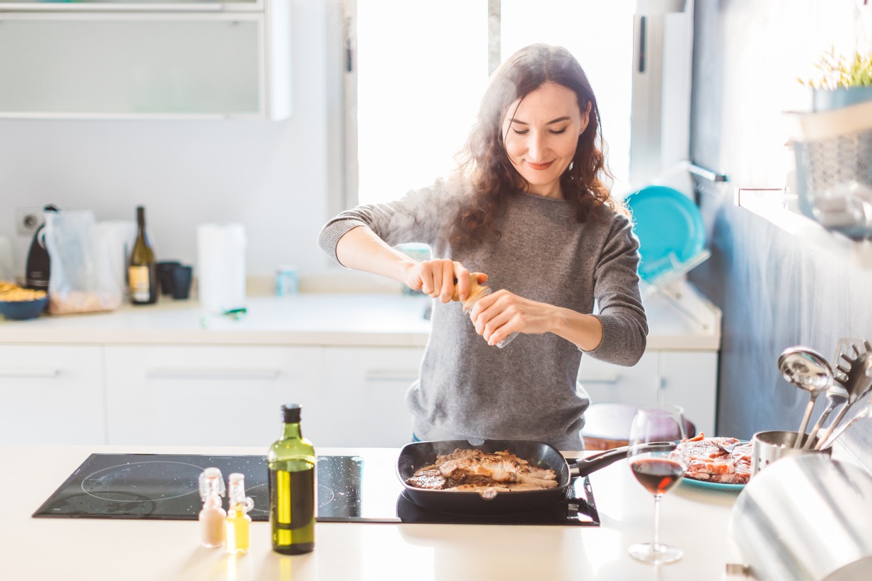 Young smiling woman cooking in the kitchen, adding pepper to the grilled meat. Healthy food concept.