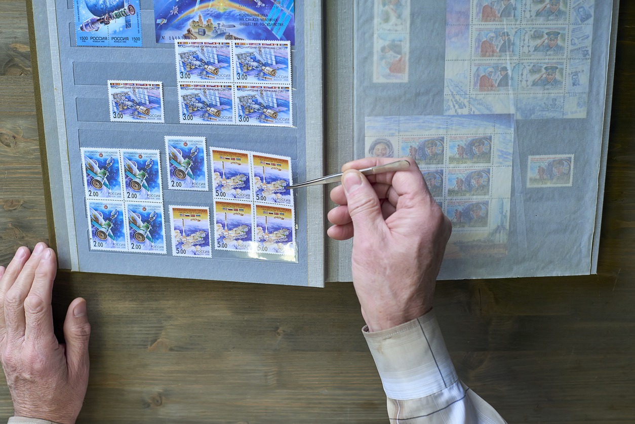 Senior man hands hold tweezers and stamp album with postage stamps collection, space theme, wooden background