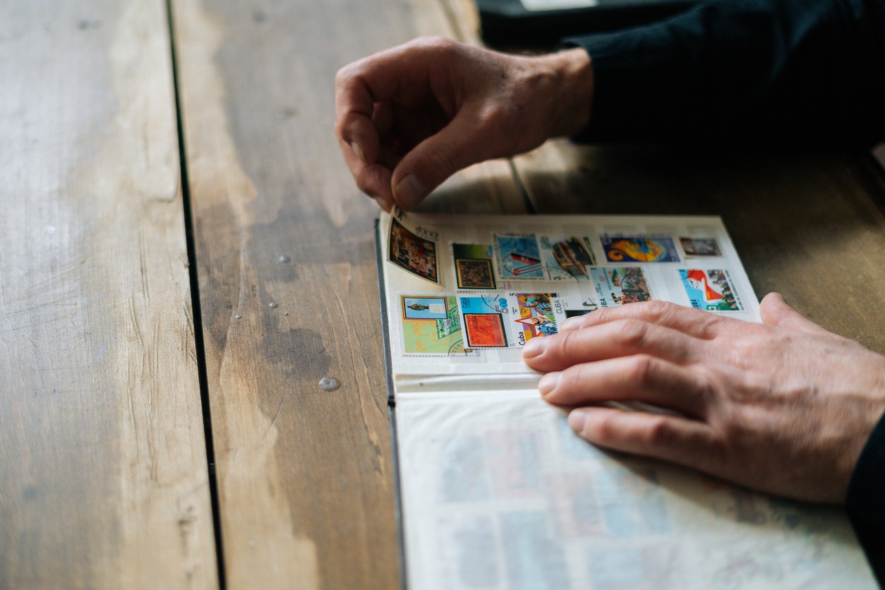 Close-up top view of unrecognizable senior adult man inserting postage stamps at collection book sitting at wooden table.