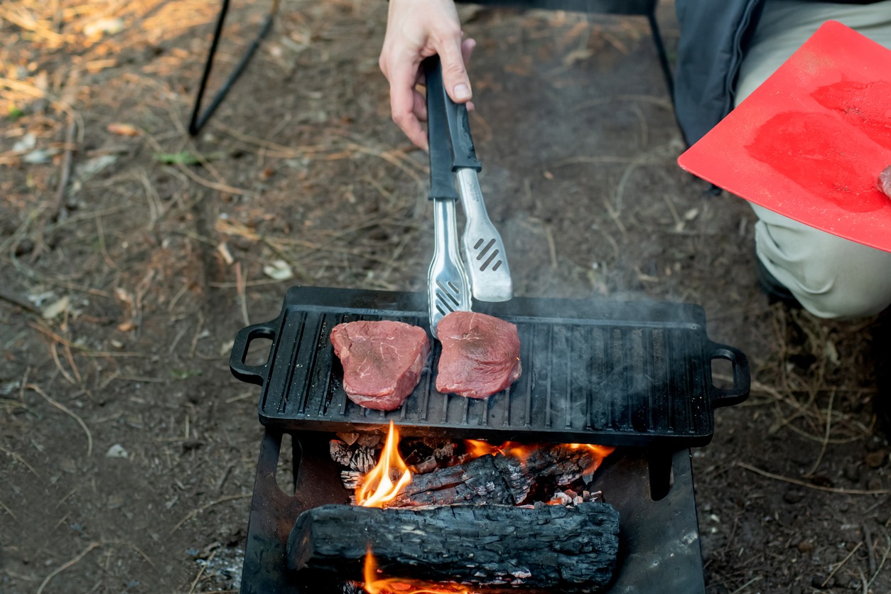 A man using tongs to cook meat at a camping site. 