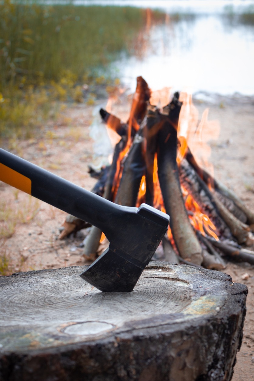 The axe is stuck in a log. Chopping wood for a fire on the background of the lake. Survival in the forest during a hike. Making a fire. A sharp axe. A woodcutter's tool. A modern axe.