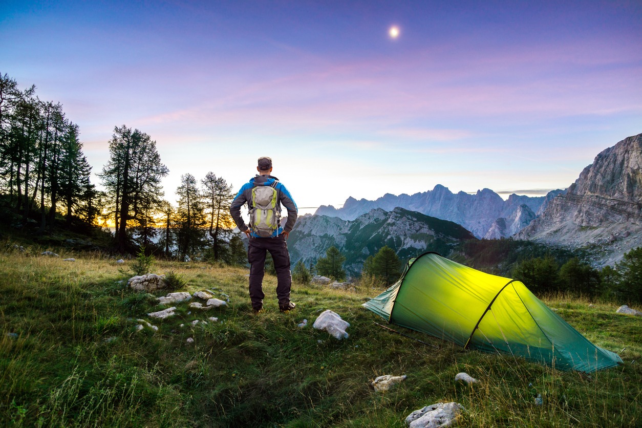 Hiker with Backpack standing on Mountain and Tent glowing under a moon night sky at sunset or sunrise twilight hour. Alps, Sleme, Triglav National Park, Slovenia.