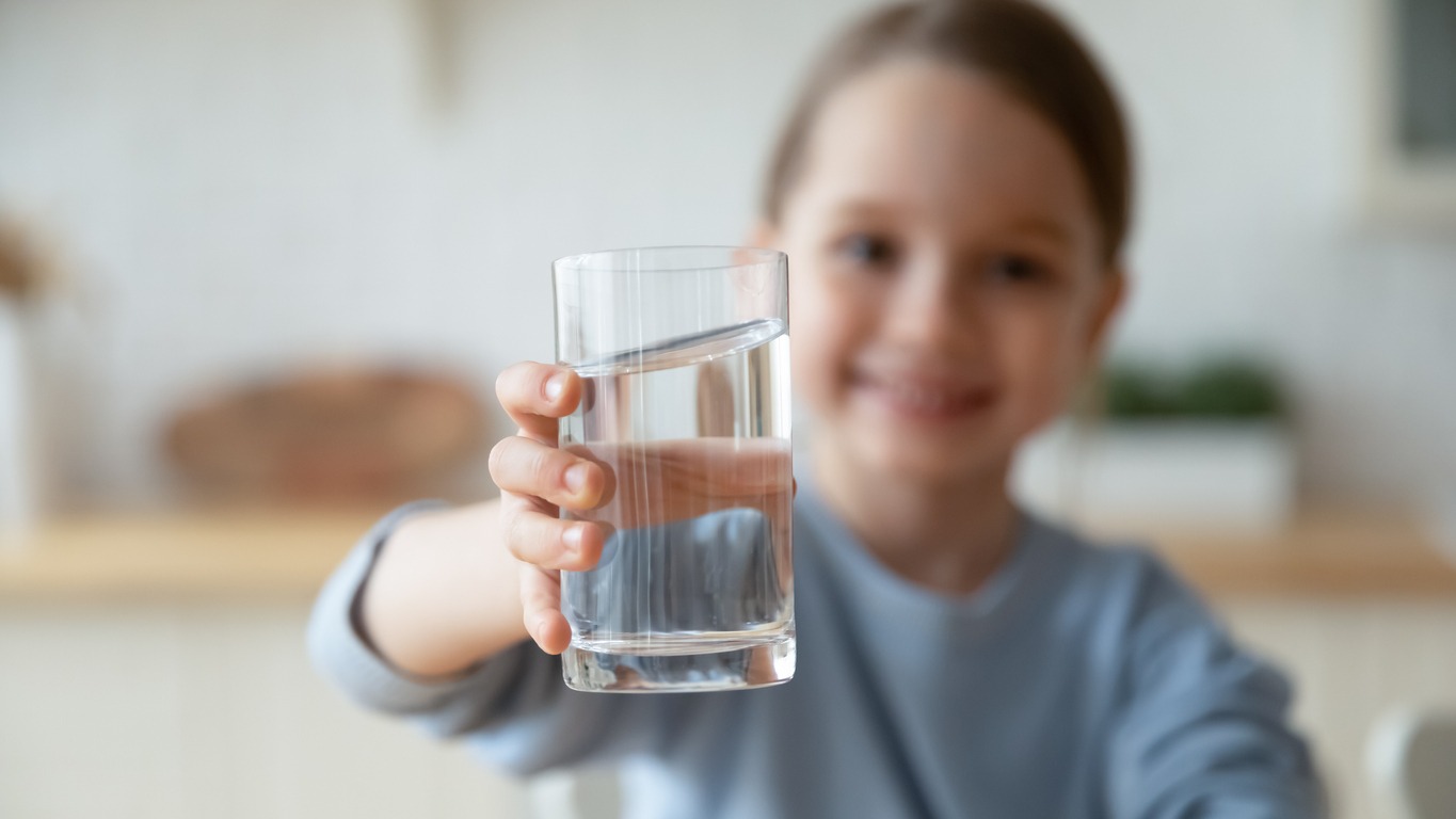 Close up smiling little girl holding a glass of pure mineral water, offering to the camera, cute pretty child kid recommending healthy lifestyle habit, drinking clean aqua for refreshment