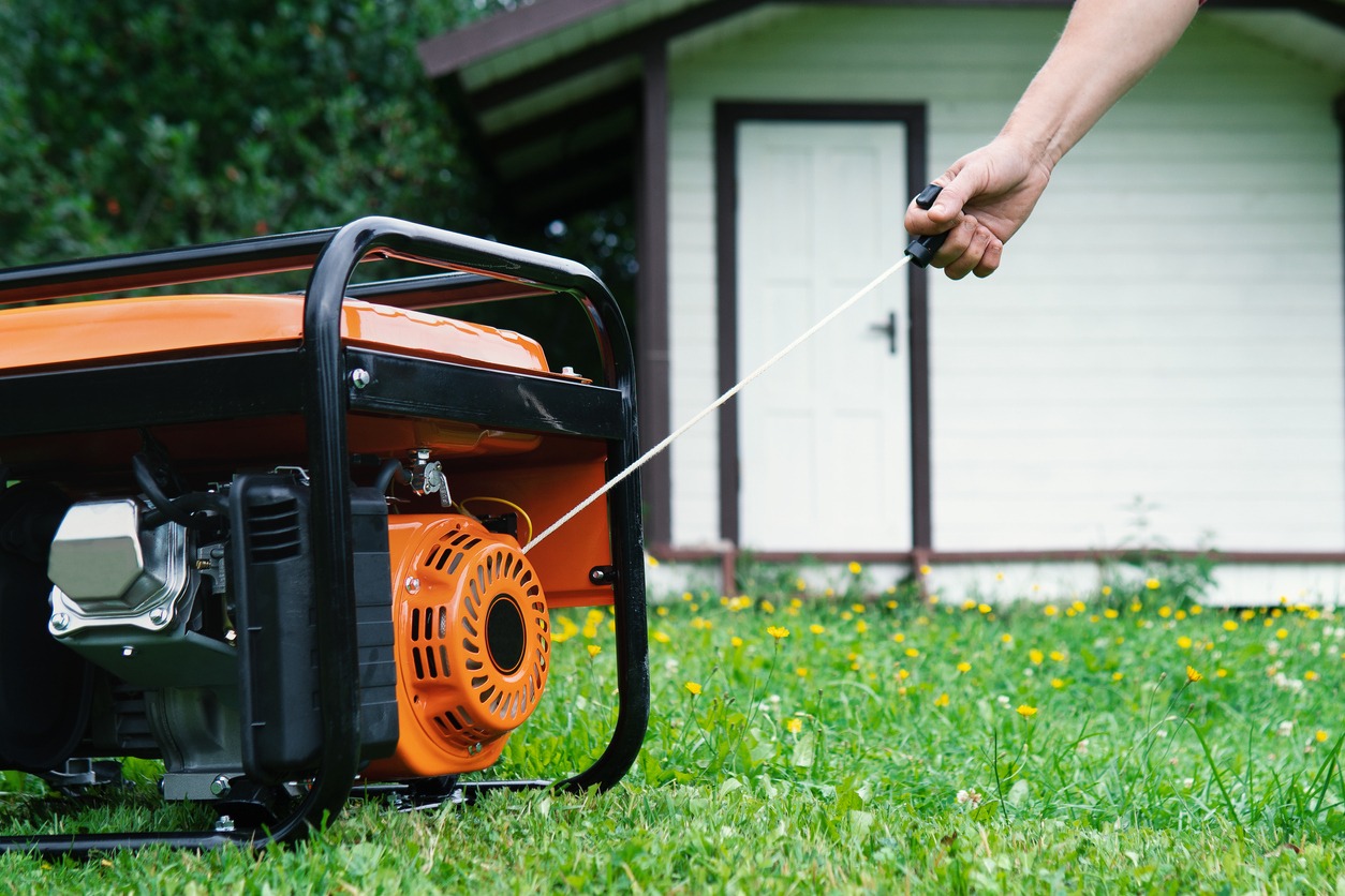 Female hand starts a portable electric generator standing on the grass in front of a summer house on a summer evening