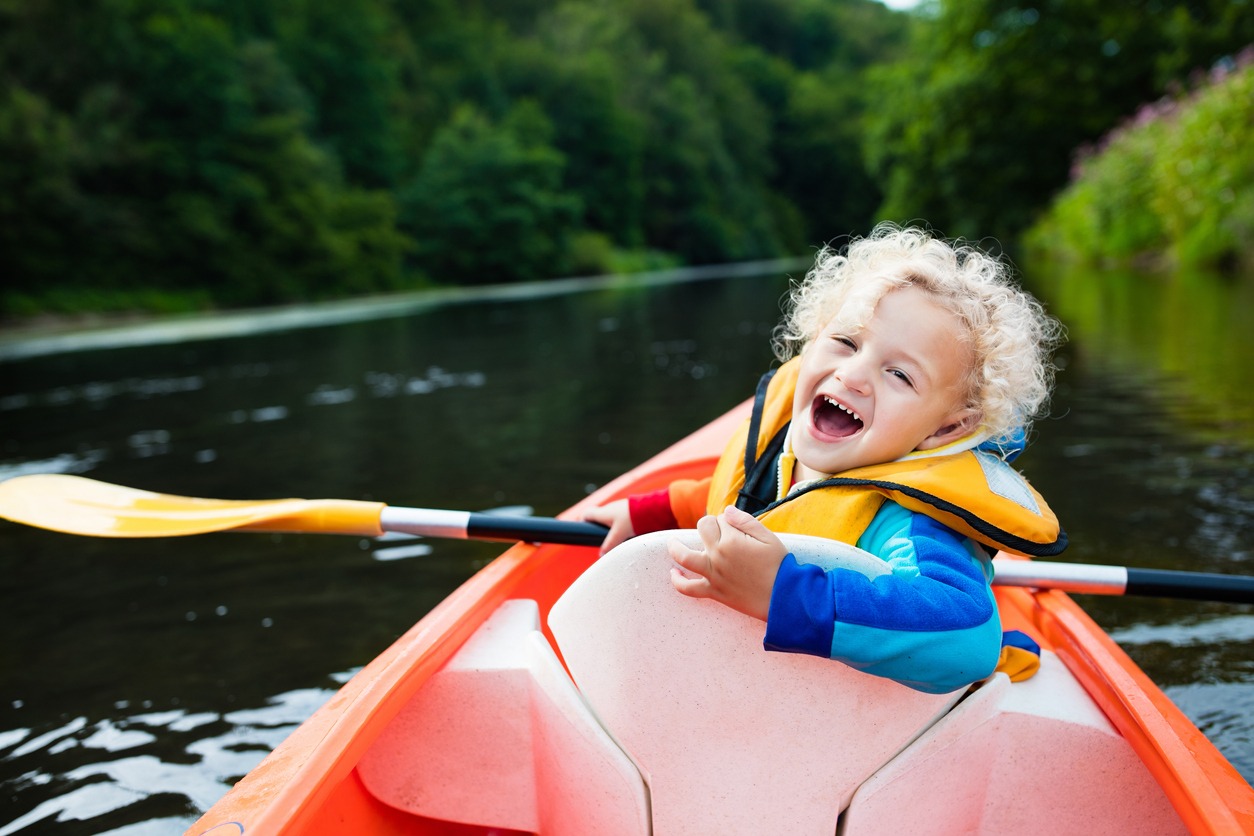 Happy kid enjoying kayak ride on beautiful river. Little curly toddler boy kayaking on hot summer day. Water sport and camping fun. Canoe for children. Funny child with vessel in a boat.