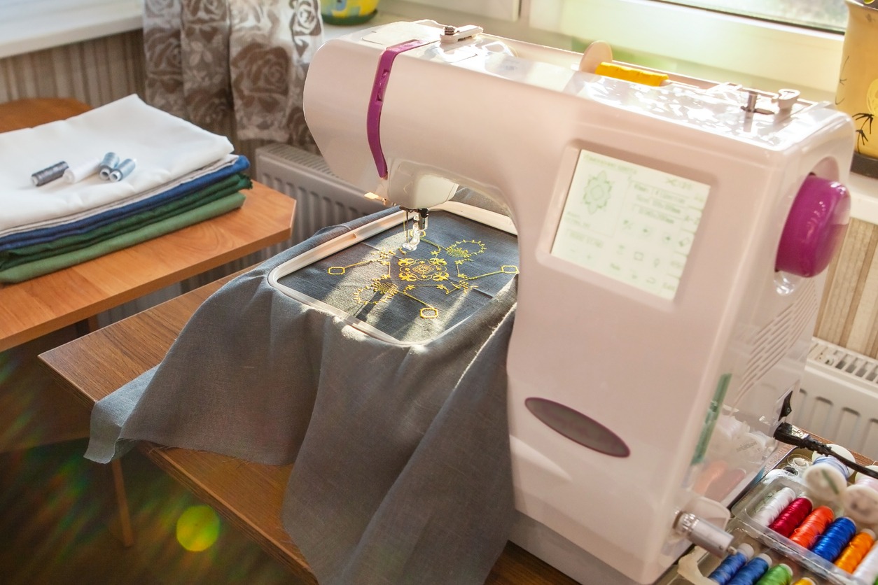 Home sewing workshop, workplace opposite the window, with a modern embroidery machine that creates a yellow pattern on a gray linen fabric. Next to the thread and fabric