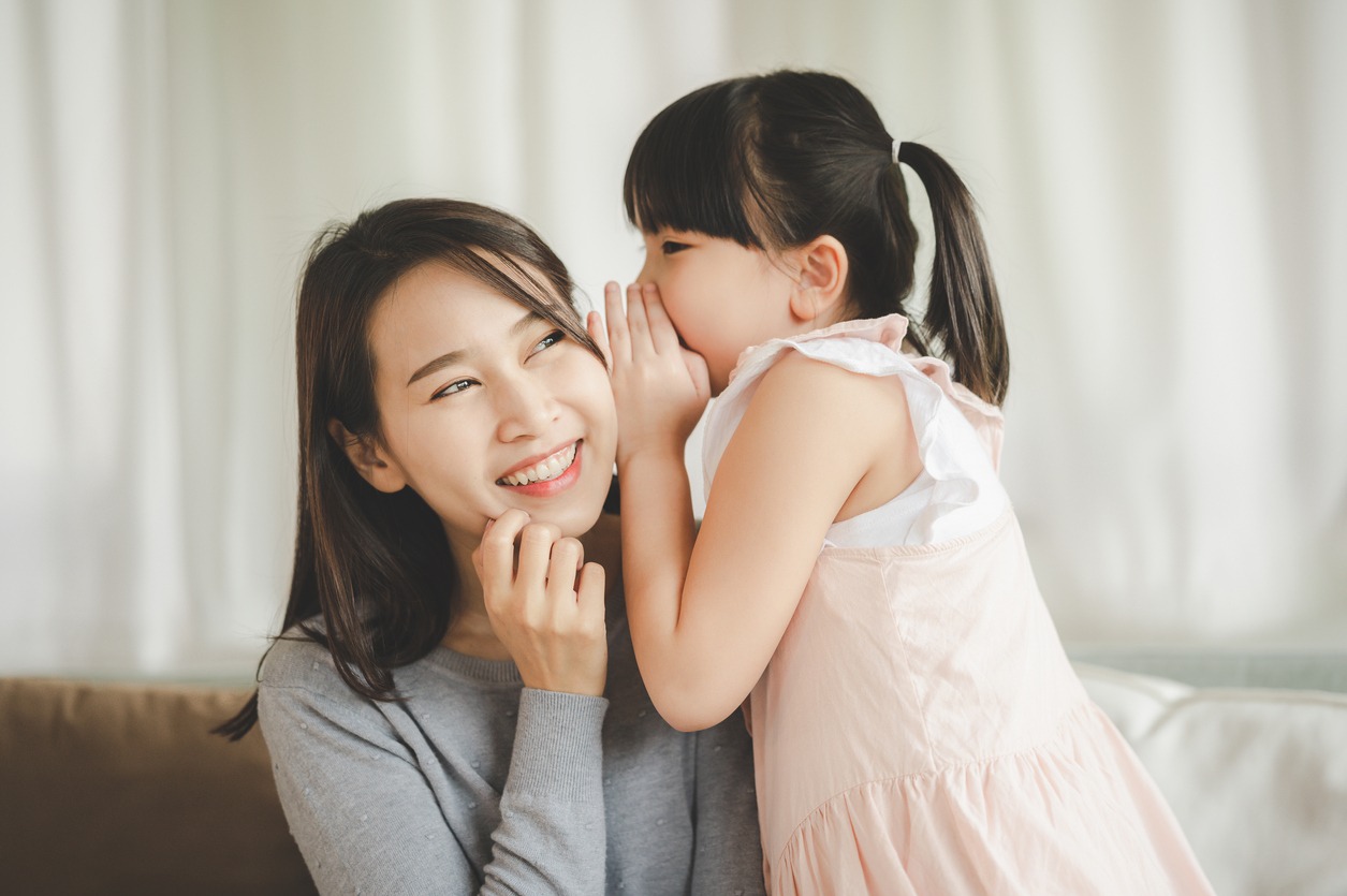 Little Asian daughter girl whispering I love you, mommy, to happy smiling mother in the living room