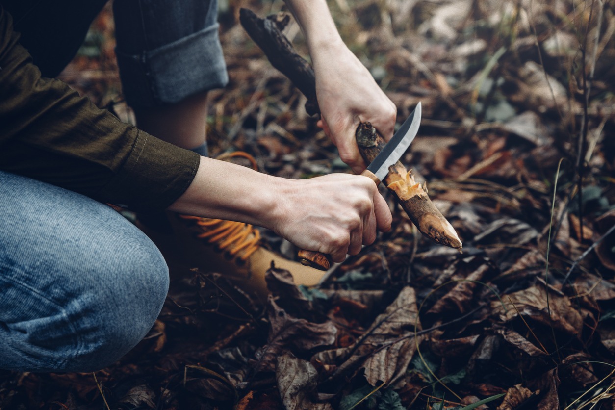 Low-angle view of mid-adult woman sharpening the end of a branch in the forest using his knife
