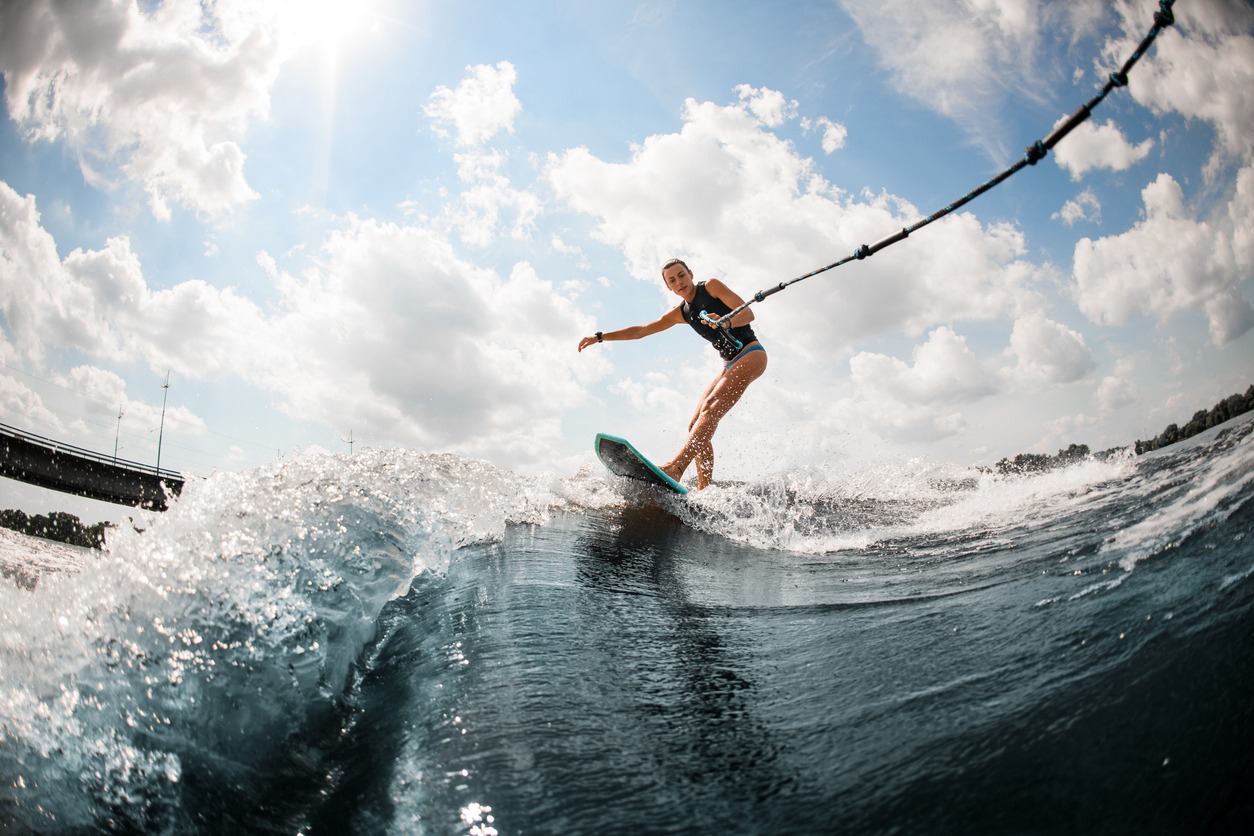 Young beautiful woman riding on the wake surf holding rope of a motorboat against blue sky with white clouds