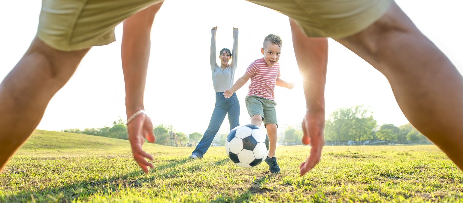 Young happy family playing football outdoors in a public park garden. mom, dad, and little child having fun together. cute kid kicking a ball in motion and scoring a goal. sport and lifestyle concept