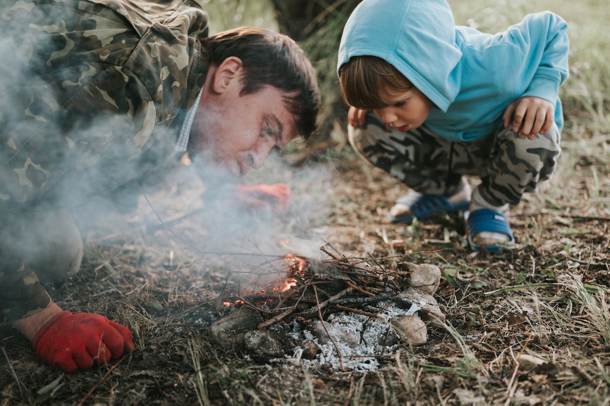little happy five-year-old kid boy and his father blowing and kindling a campfire in the village or forest on nature outdoors on a summer evening. authentic countryside and children's rustic life