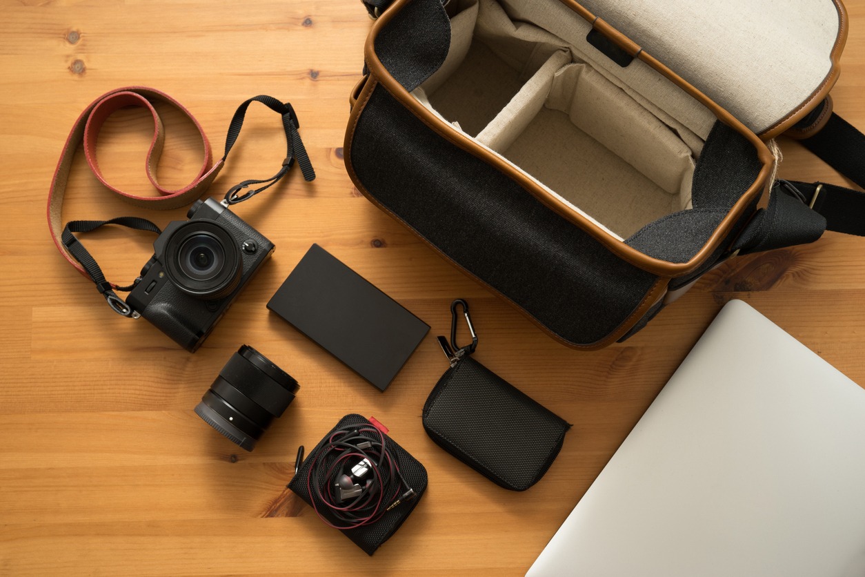 Black digital camera with orange leather strap on wooden table with navy bag, laptop, power bank, lens, and earphone