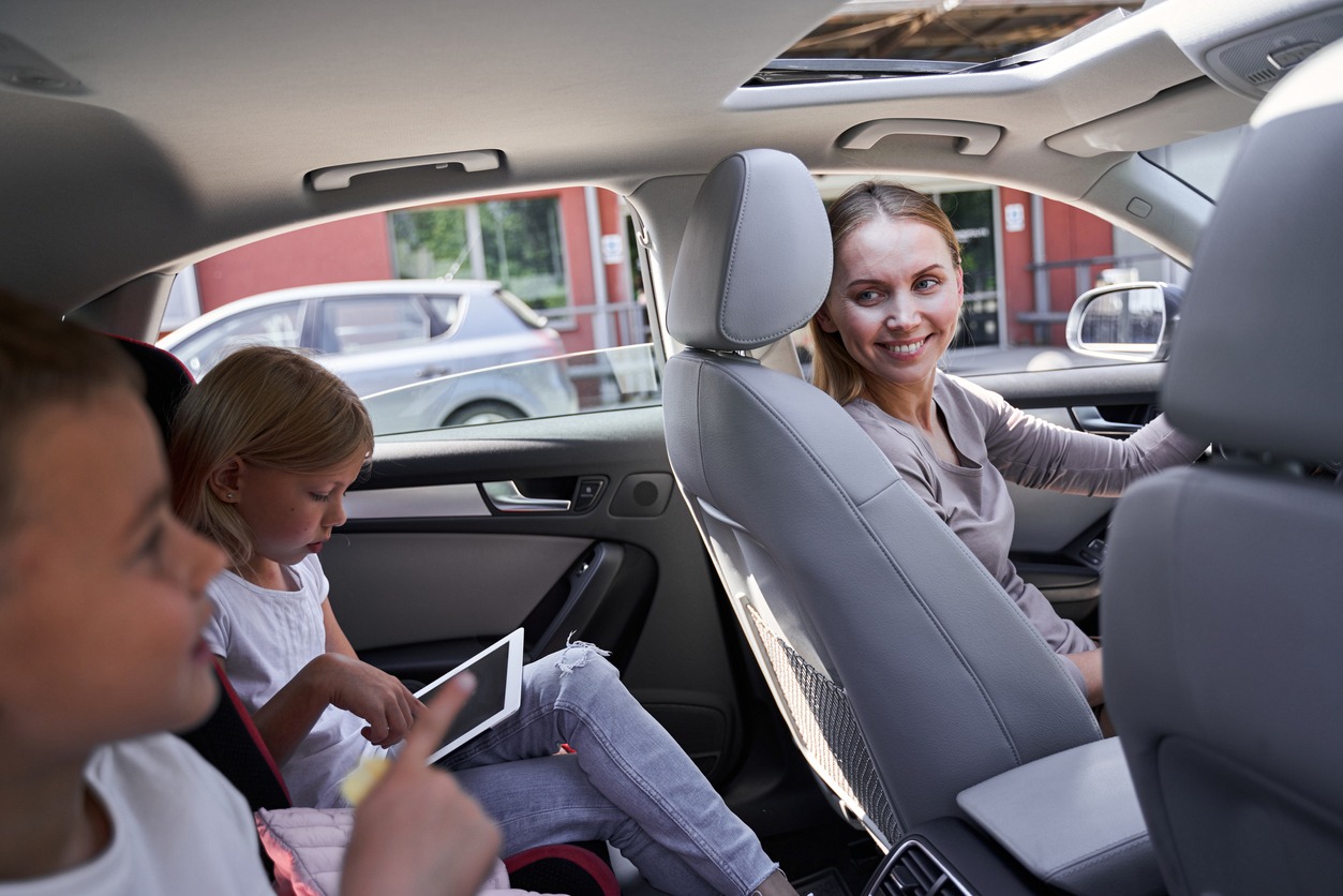 Cheery attractive mother driving car and looking at son and daughter with smartphone and tablet