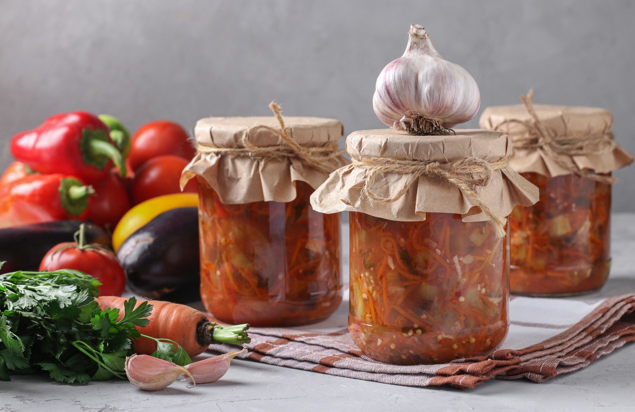 Pickled salad with eggplant, carrot, peppers, garlic, and tomatoes in glass jars on light gray background