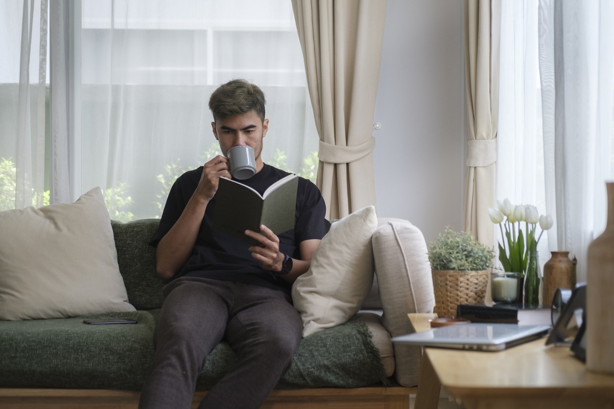 Positive young man reading a book and drinking coffee while sitting on the sofa at home