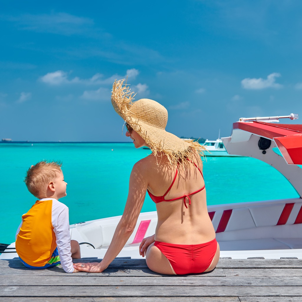 Three year old toddler boy with mother sitting on wooden jetty with boat. Summer family vacation at Maldives.