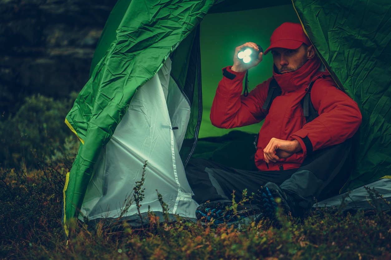 A man using a flashlight sitting outside a tent.