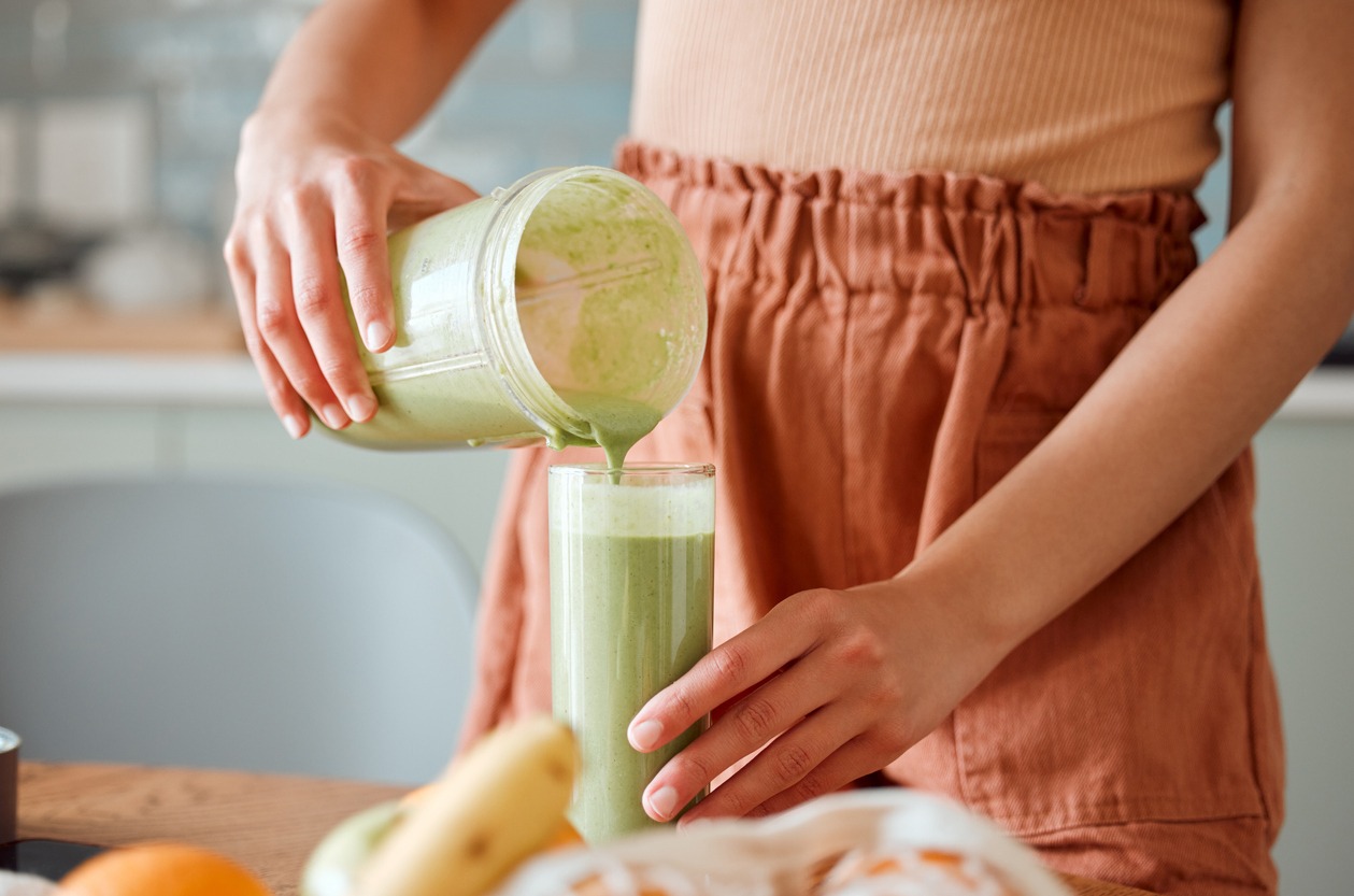 Woman pouring healthy smoothie in a glass from a blender jar on a counter for detox Female making fresh green fruit juice in her kitchen with vegetables and consumables for a fit lifestyle